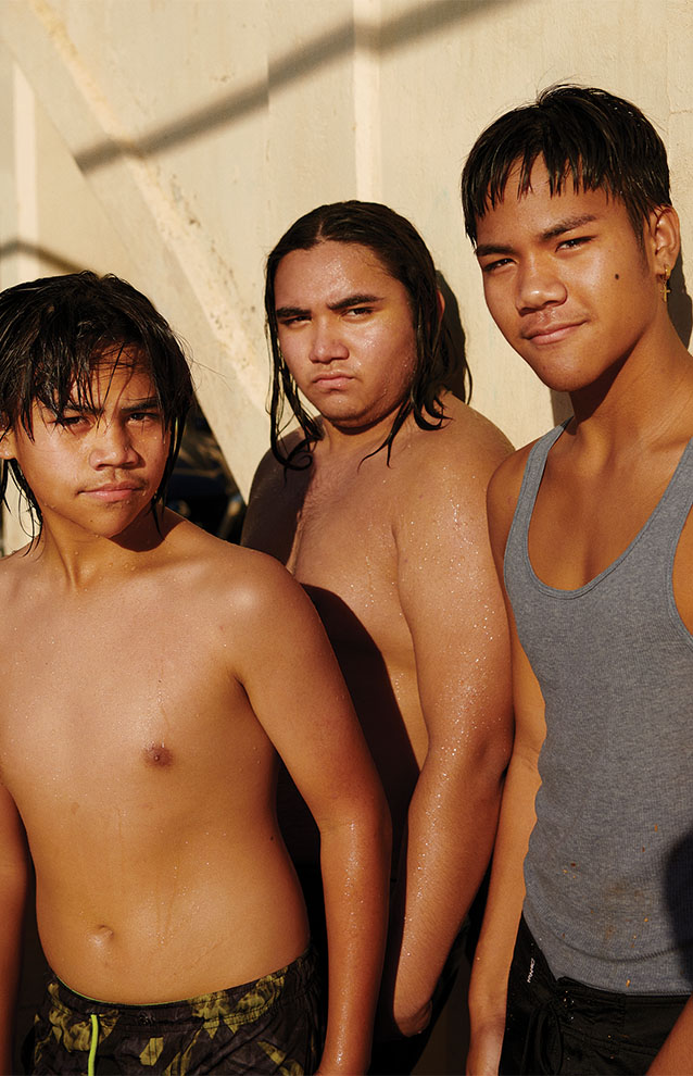 three young men in swim trunks and a tank top stand against the bridge in the sun. 