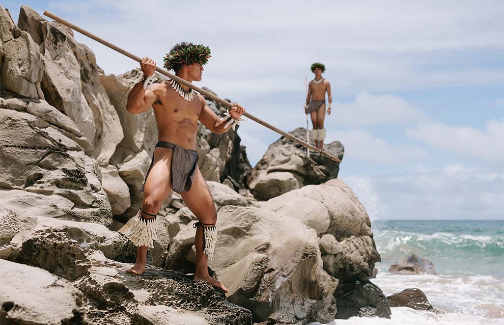 Two people in traditional attire stand on rocks by the ocean, one holding a spear, both wearing garlands on their heads.