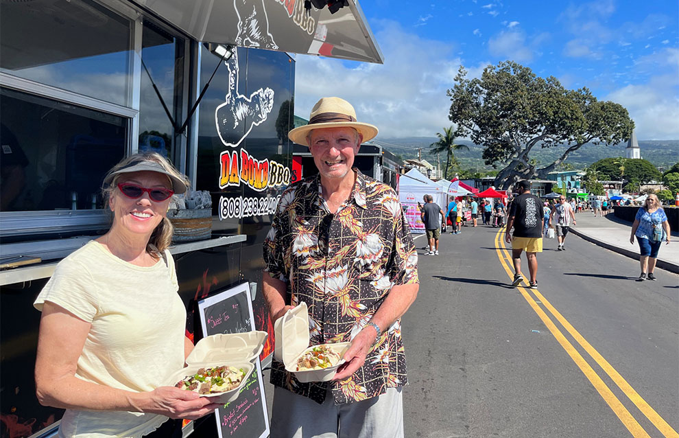 two people standing outside beside a black food truck, holding food